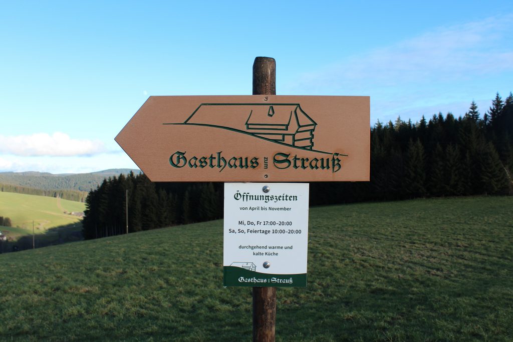 A directional sign on a post sporting the logo of the Gasthaus zum Strauss and its opening times, backed by forest and hills.